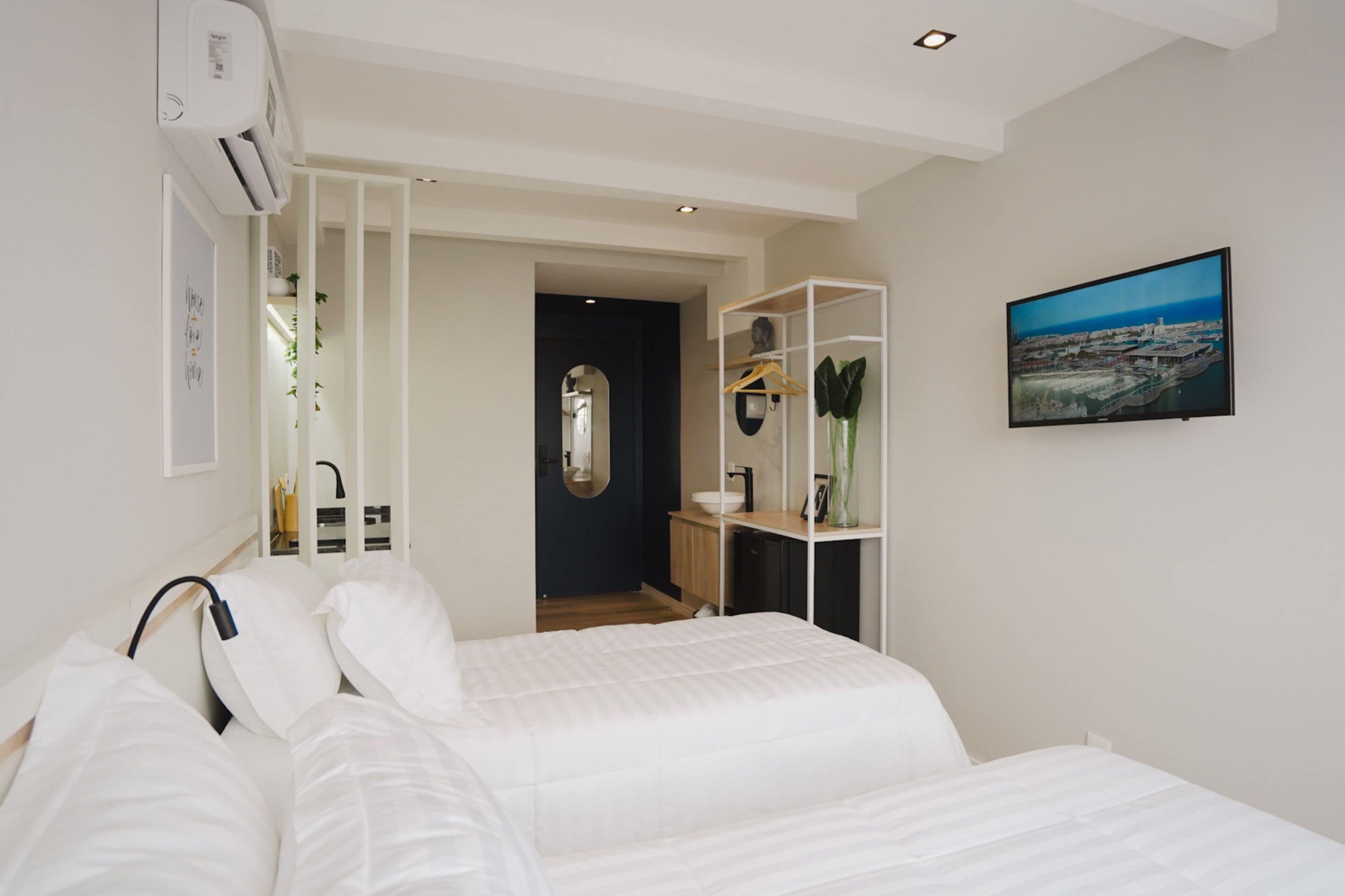 Habitación de hotel moderna con una cama bien tendida, una mesita de noche, un espejo, una repisa con adornos y una pintura de paisaje urbano en la pared.