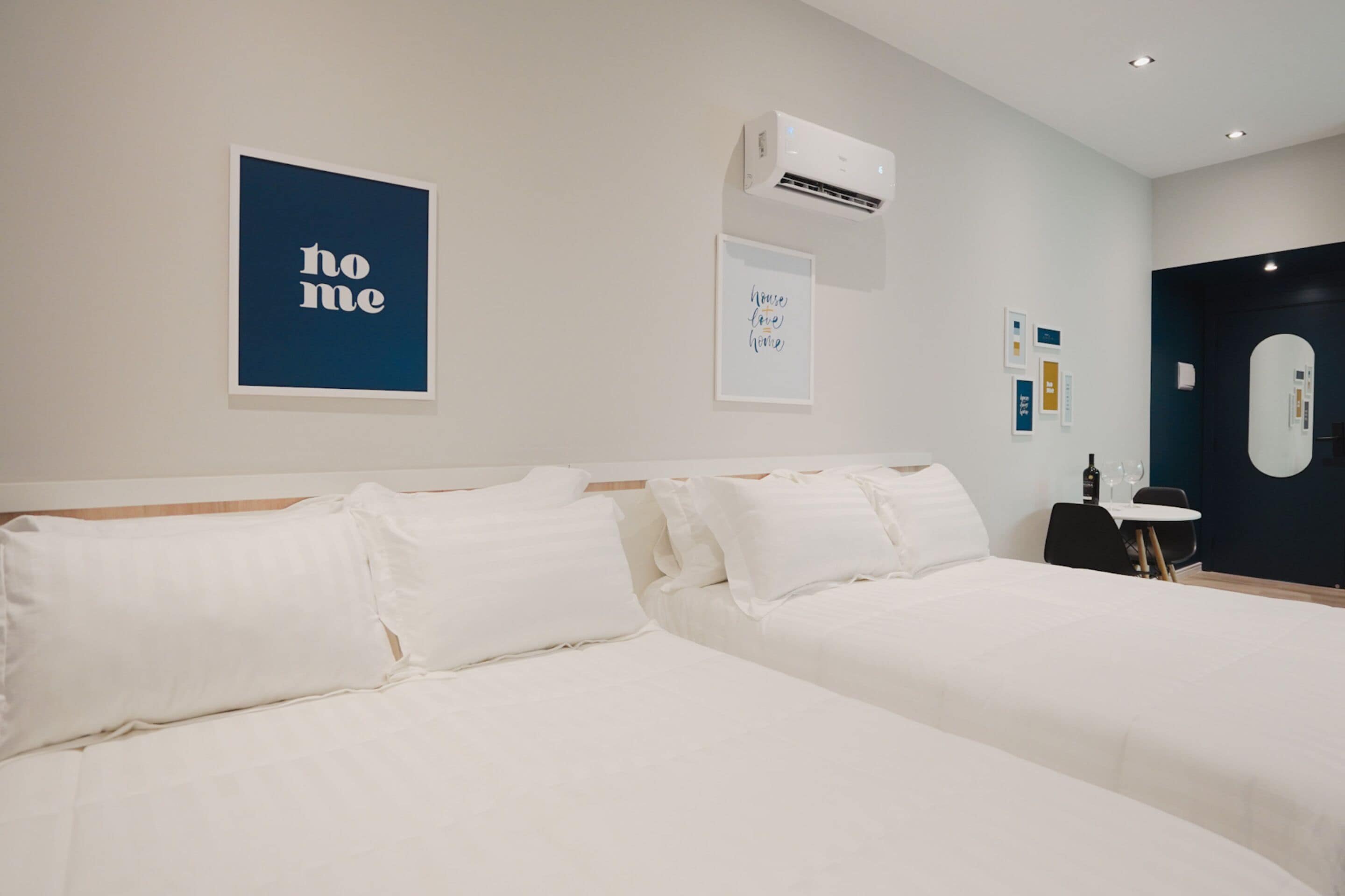 Quarto de hotel com duas camas, uma mesinha com uma cadeira e obras de arte penduradas na parede.