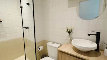 Uma casa de banho moderna com cabine de duche em vidro, sanita branca, lavatório com torneira preta e espelho na parede.