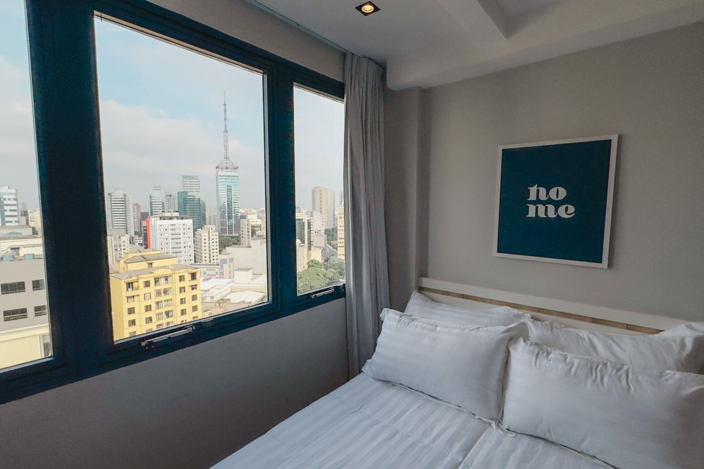 A hotel room with a large window offering a city view, a neatly made bed, and a 'no me' sign on the wall.