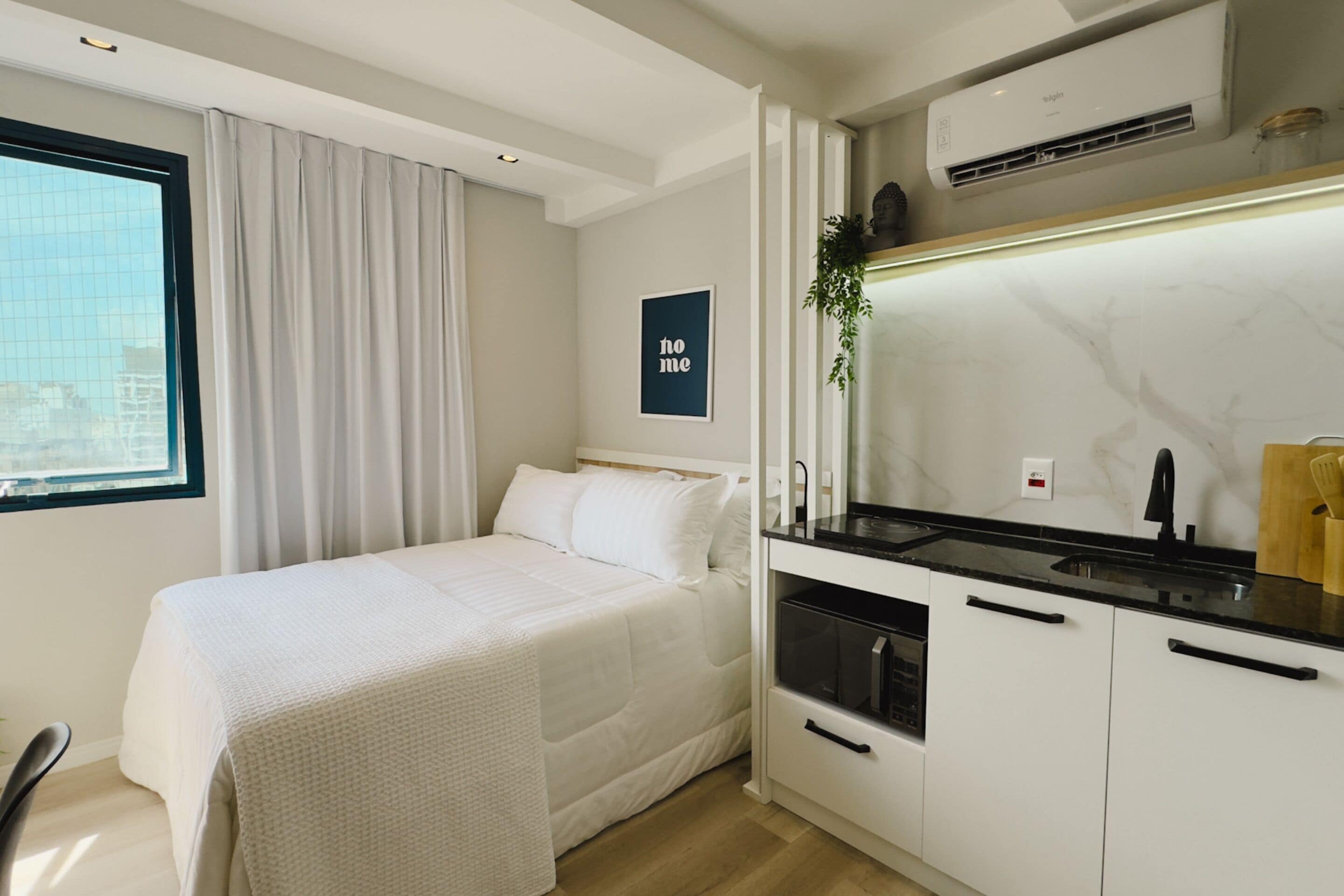 Een moderne hotelkamer met een bed, een kitchenette met een spoelbak en oven, een raam met gordijnen en uitzicht op de stad.