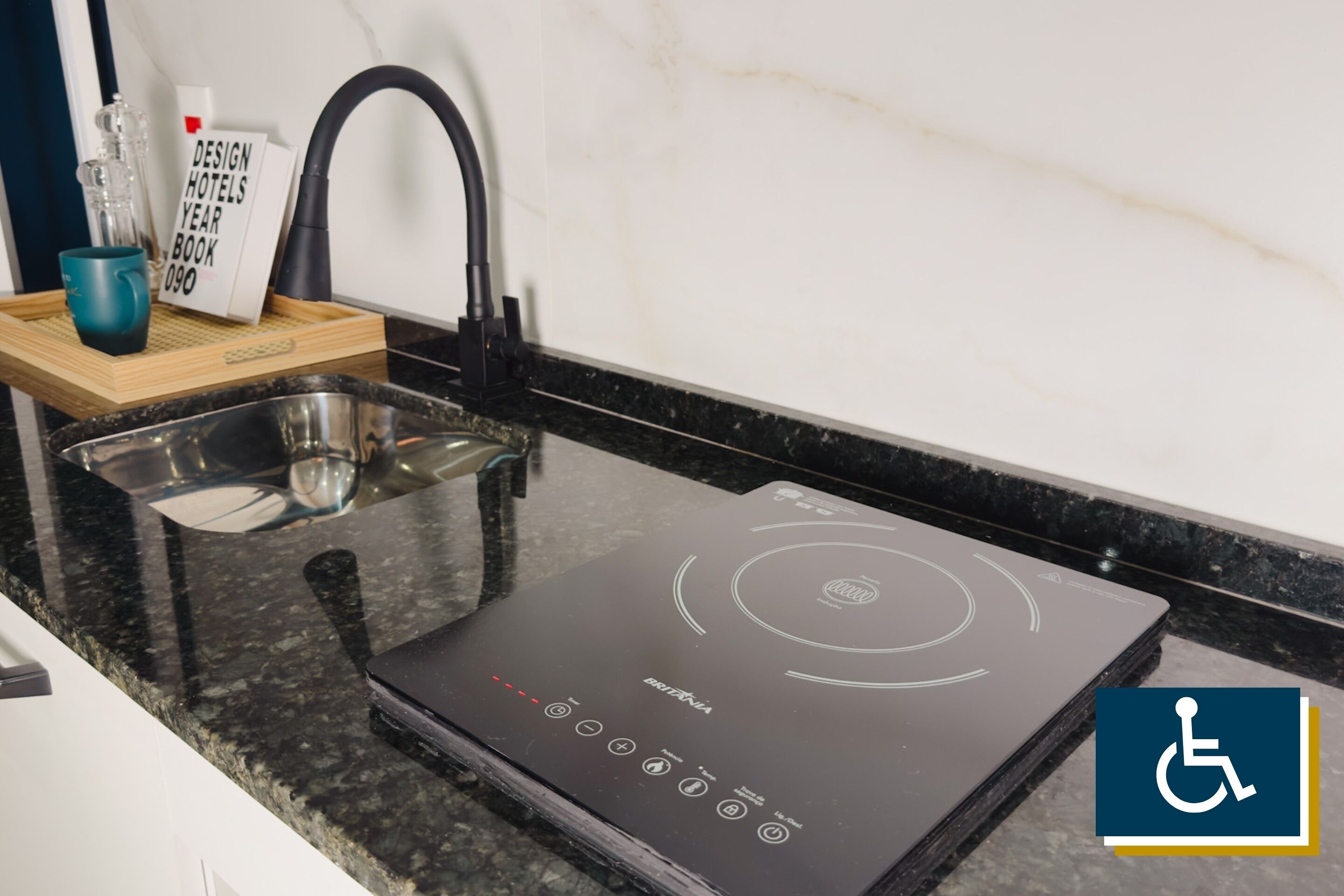 Une cuisine dotée d’une plaque de cuisson à induction noire, d’un évier en acier inoxydable et d’un plateau en bois sur lequel se trouvent une tasse bleue et un livre intitulé « DESIGN HOTELS YEAR BOOK 090 ».