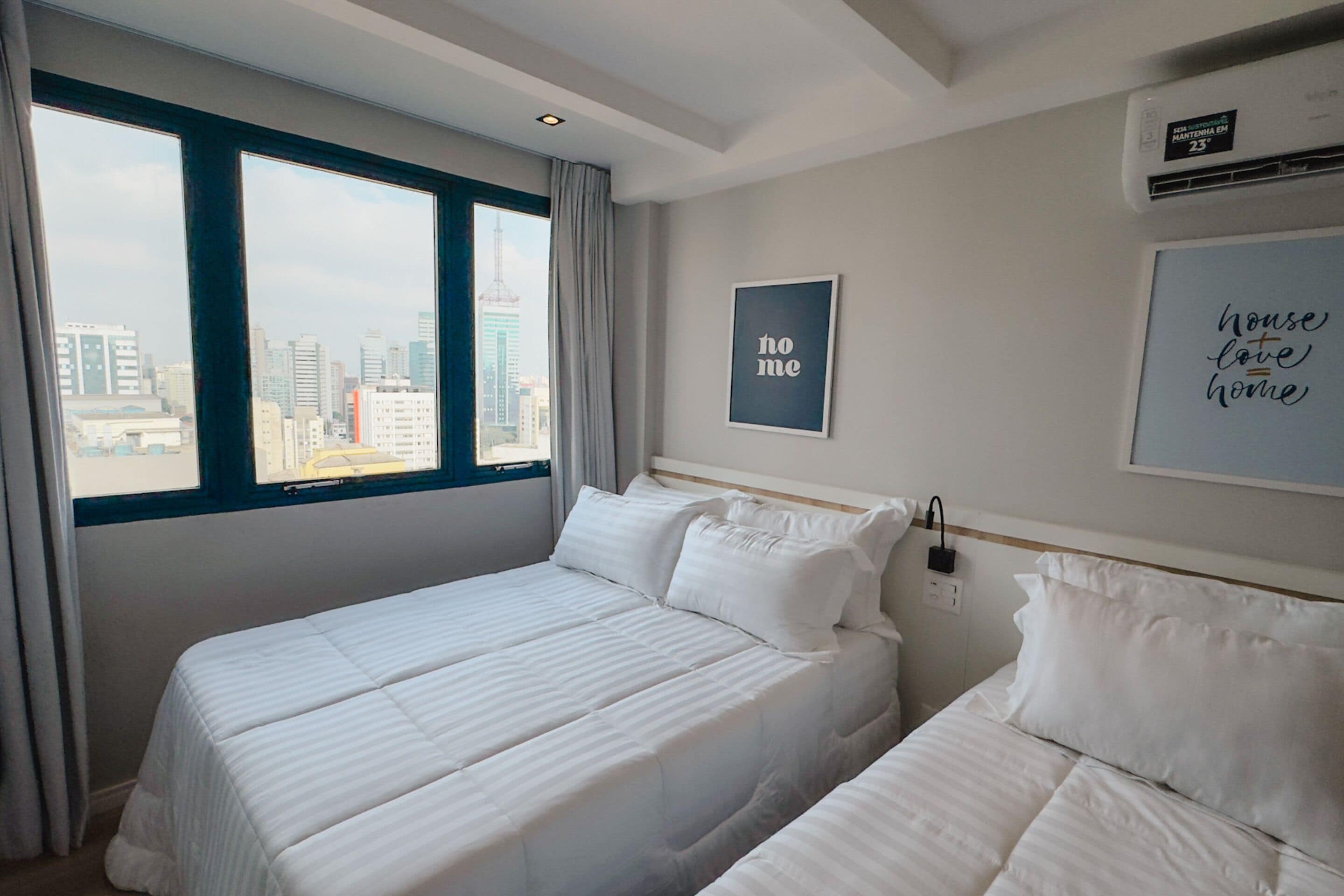 Hotellihuone, jossa on kaksi sänkyä, näkymä kaupunkiin ja seinällä oleva kehystetty kyltti, jossa lukee ”koti”.