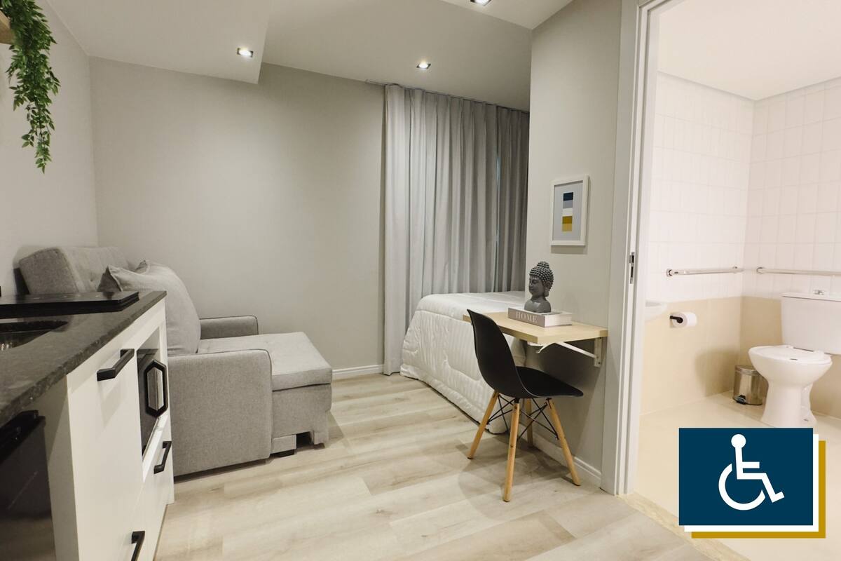 En modern lägenhet med ett litet kök, en grå soffa, ett litet skrivbord med en stol, en säng och ett badrum med toalett.