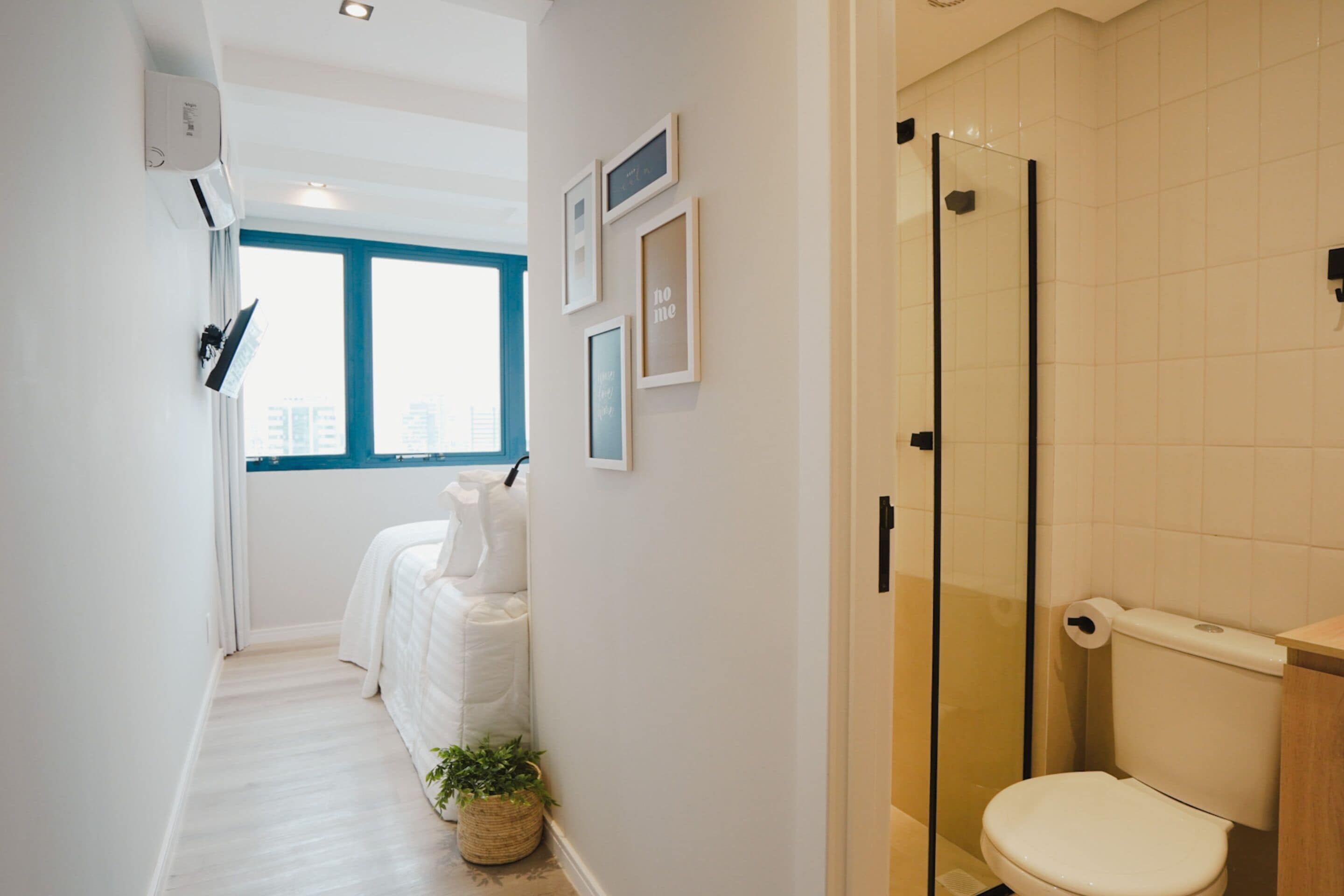 A modern bathroom with a glass shower enclosure, a white toilet, and a bed with white bedding in the adjacent room.