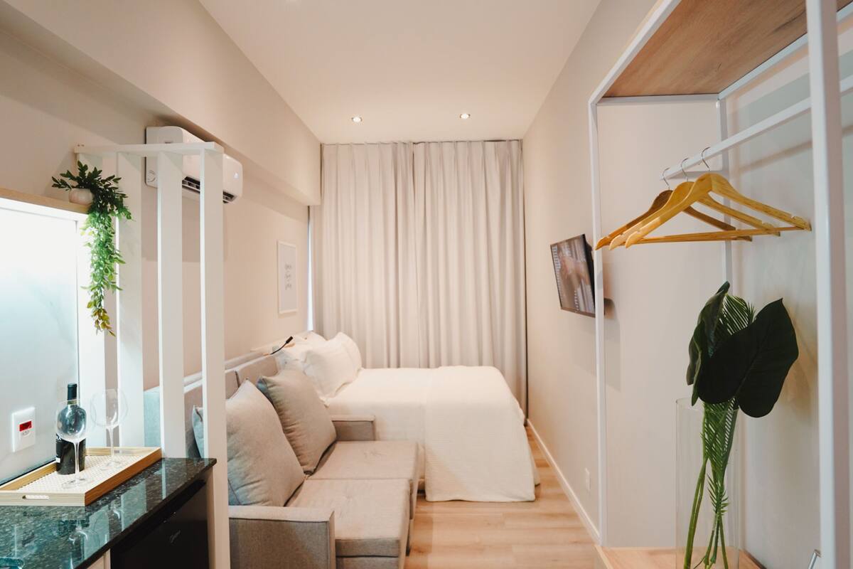 Ett modernt hotellrum med en säng, en soffa, ett litet bord med en flaska och glas, en växt och en garderob med galgar.
