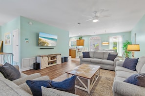 A living room with a sofa, coffee table, and TV. There is a kitchen in the background.