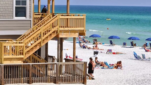 A beach house with a wooden deck, a lifeguard tower, and a sandy beach with umbrellas and people.