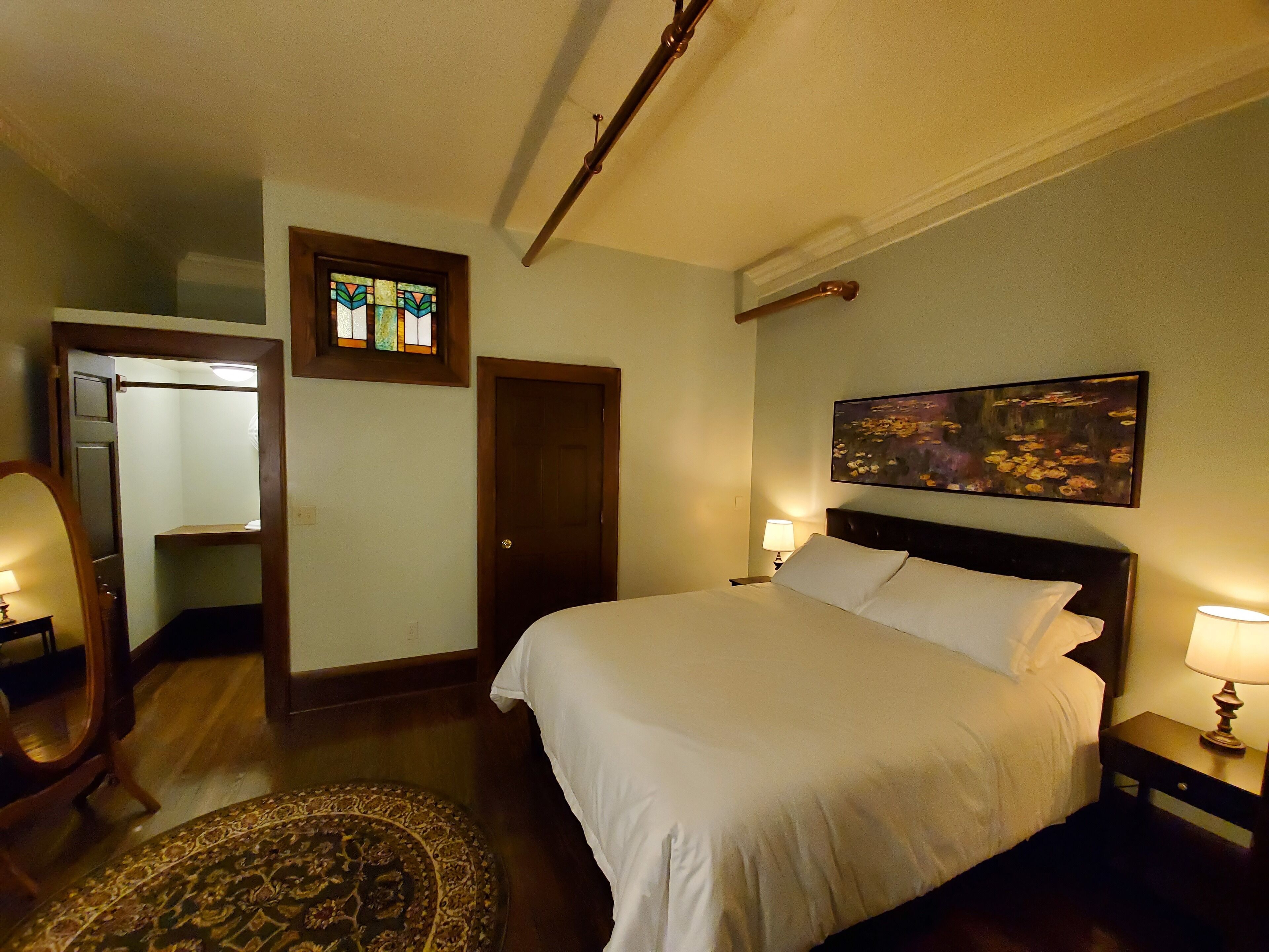 A hotel room with a bed, a nightstand, a lamp, a painting on the wall, a door, and a mirror.
