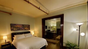 Quarto de hotel com uma cama grande, luminárias de cabeceira, um quadro, um espelho, uma escrivaninha com cadeira e uma planta em vaso.