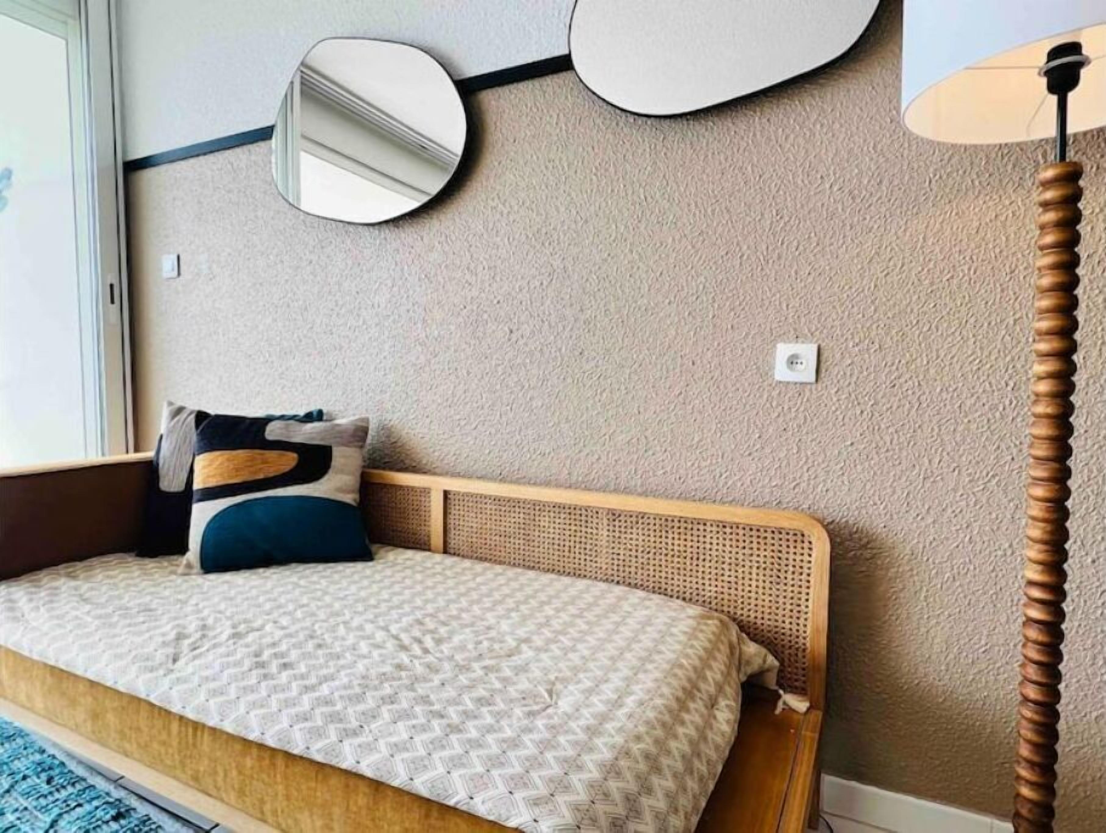 Een slaapkamer met een houten hoofdbord, een bed met een patroondessin, één kussen en een ronde spiegel aan de muur.