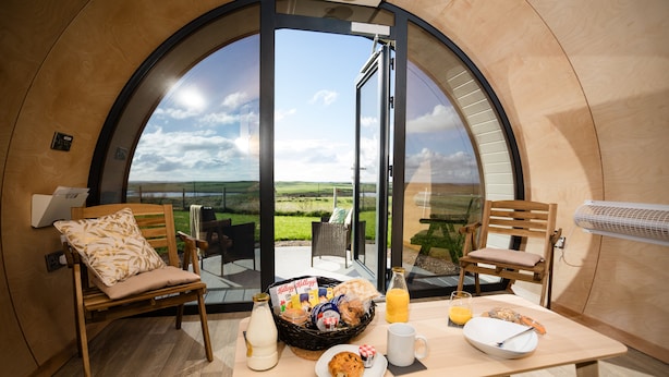 Une table de petit-déjeuner est dressée dans une pièce circulaire et moderne, dotée d’une grande fenêtre, et comprend une table avec des aliments pour le petit-déjeuner et deux chaises.