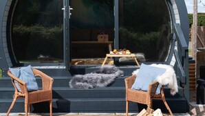 Un bâtiment moderne de forme circulaire, doté d'une grande porte en verre, de chaises en osier avec des coussins blancs, d'une petite table où sont disposés des aliments et d'un foyer.