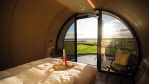 Une chambre à coucher avec un grand lit, une table de chevet sur laquelle se trouvent une bouteille et des verres, une chaise, et une vue sur un paysage verdoyant par une grande fenêtre en arc de cercle.