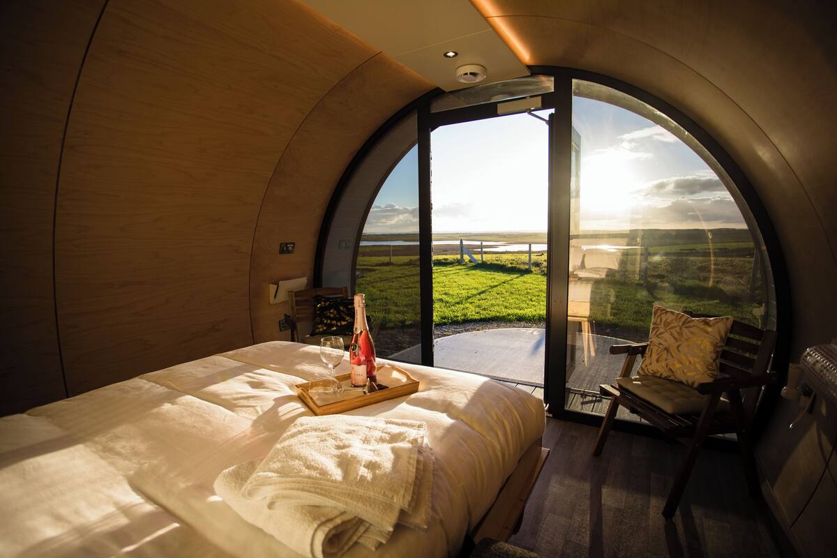 Een slaapkamer met een groot bed, een nachtkastje met een fles en glazen, een stoel en uitzicht op een grasrijk landschap door een groot, rondboogvormig raam.