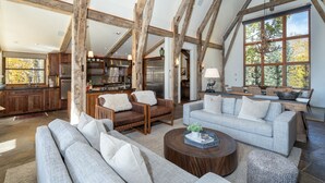 A spacious living area with a high ceiling and exposed wooden beams, a large window with a view of trees, and a modern kitchen in the background. - Mountain House by Exceptional Stays (Mountain Village)