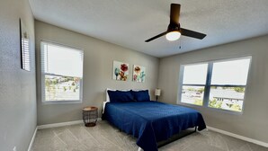 A bedroom with a bed, two windows, a ceiling fan, and artwork on the wall.