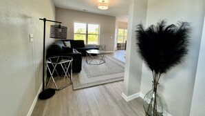 A spacious living room with a dark sofa, a glass coffee table, and a large window providing natural light.
