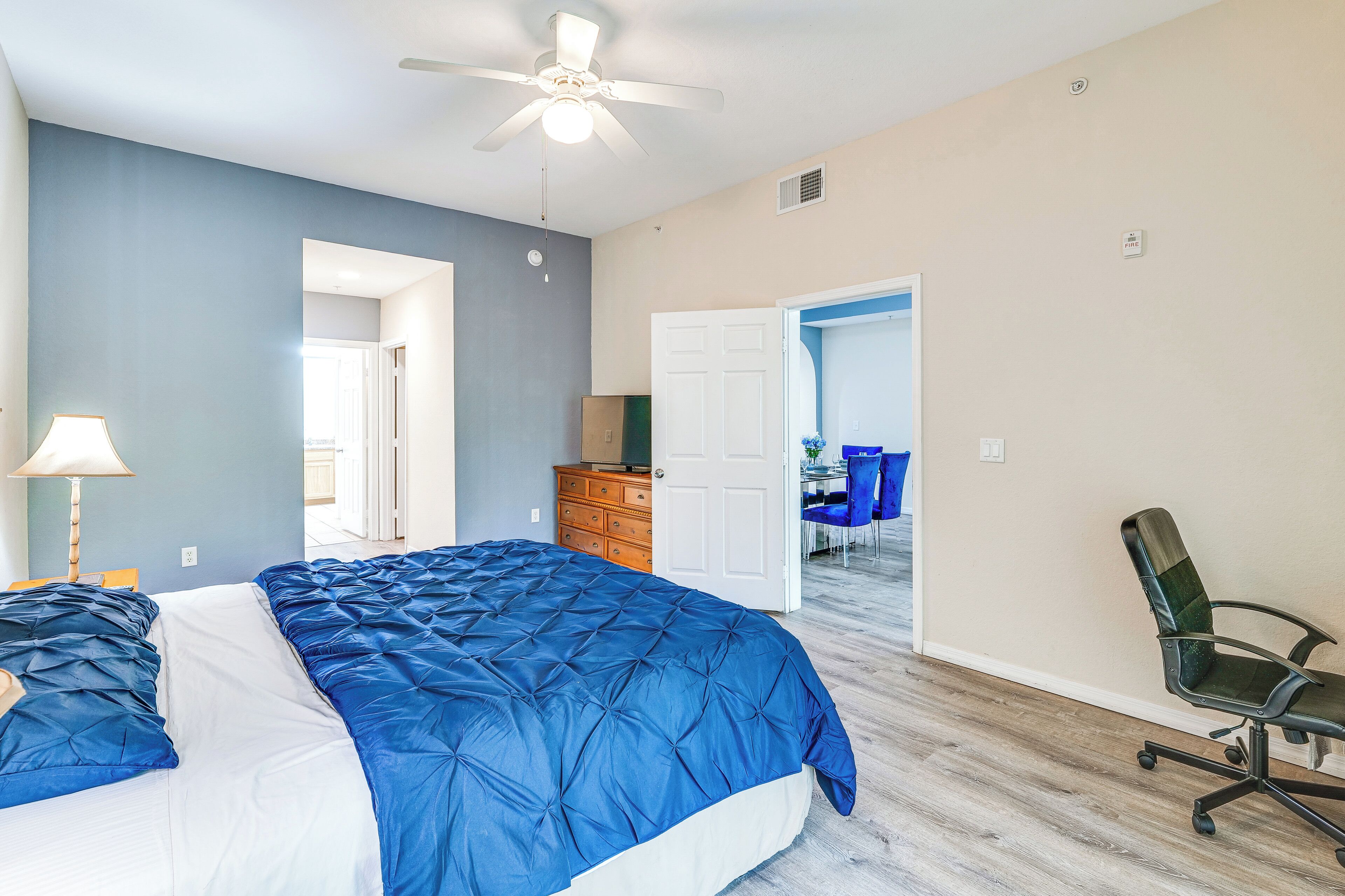 A bedroom with a bed, a desk, a chair, a dresser, and a door leading to another room.