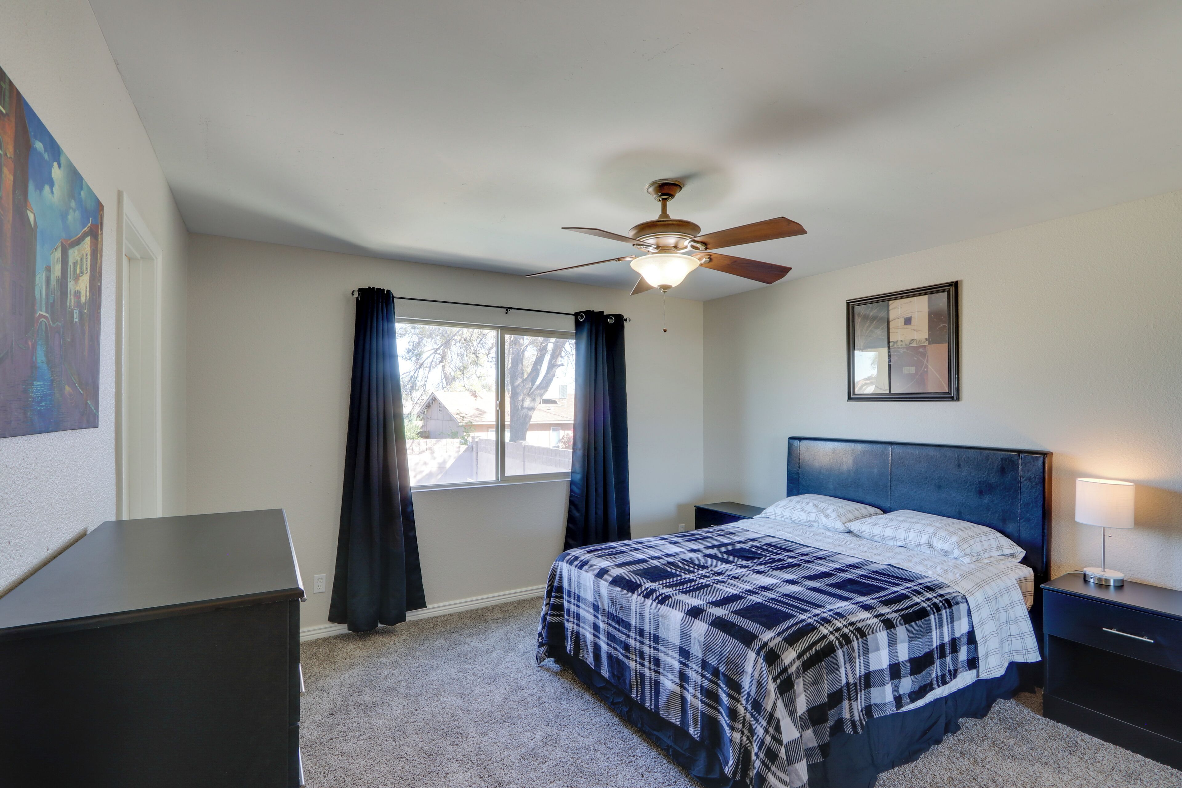 A bedroom with a bed, a nightstand, a ceiling fan, a window with curtains, and a painting on the wall.