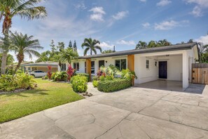 Une maison moderne à deux étages, dotée d’un patio couvert, d’une voiture garée dans l’allée et d’un aménagement paysager luxuriant de style tropical.