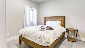 A bedroom with a wooden bed, white bedding, a round side table, and a window with white curtains. - Unique Grand Junction Guest Suite w/ Gas Fire Pit! (Grand Junction)