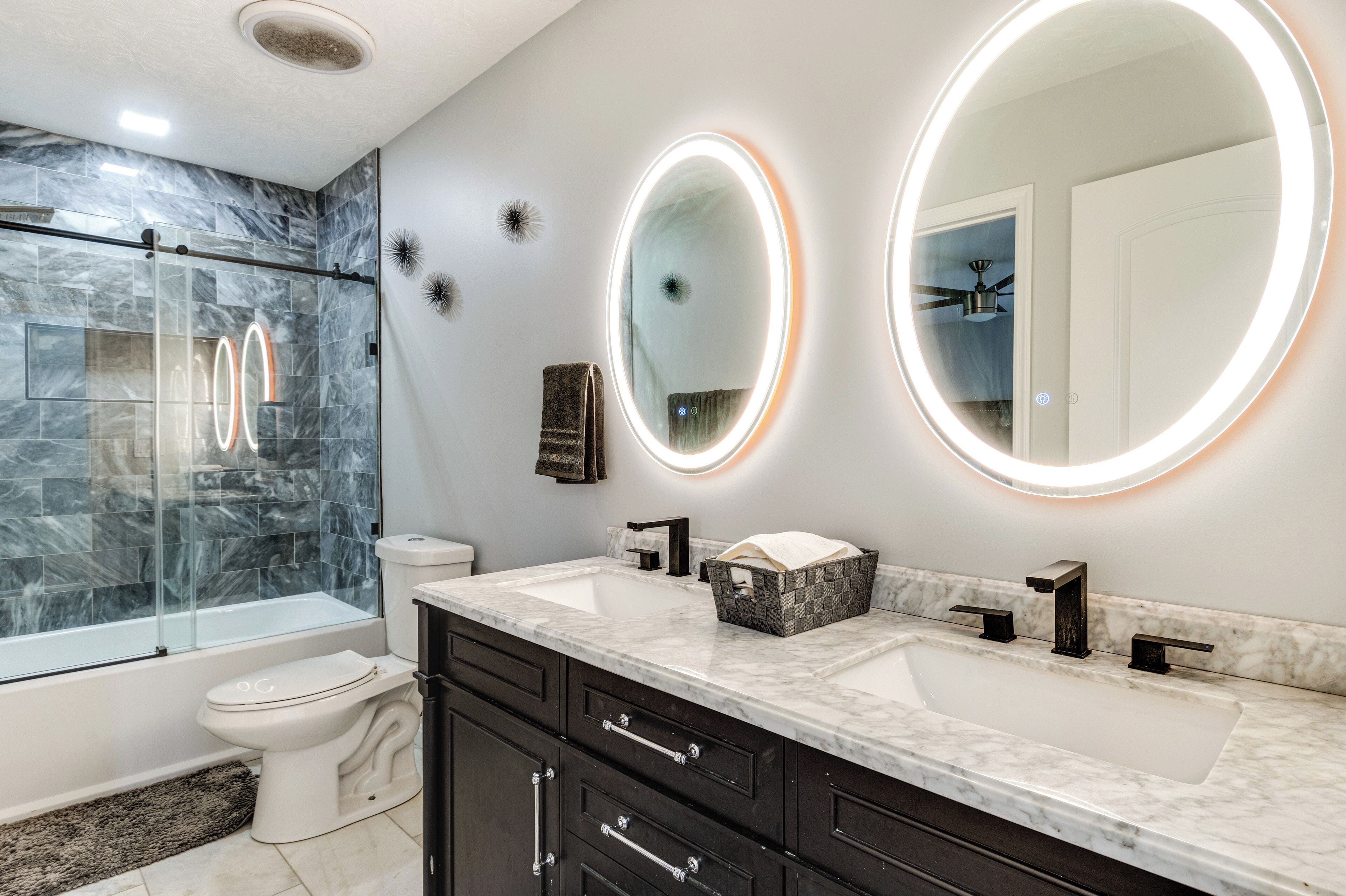 A modern bathroom with a glass-enclosed shower, a toilet, a sink with a marble countertop, and two wall-mounted mirrors with illuminated edges.