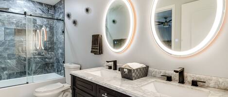 A modern bathroom with a glass-enclosed shower, a toilet, a sink with a marble countertop, and two wall-mounted mirrors with illuminated edges.