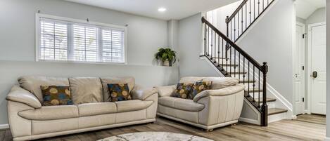 A living room with a beige sectional sofa, a round marble rug, a staircase with a wooden handrail, and a window with blinds.