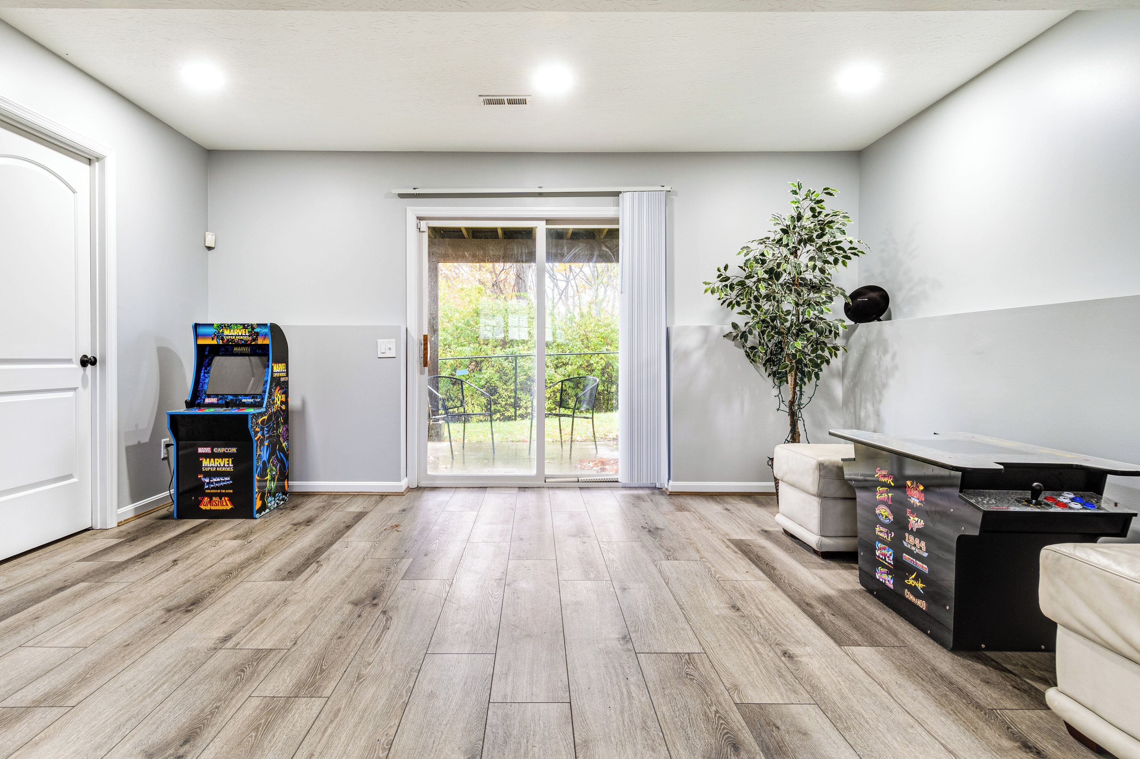 A spacious room with a wooden floor, a sliding glass door leading to a balcony, a small arcade machine, and a pool table.