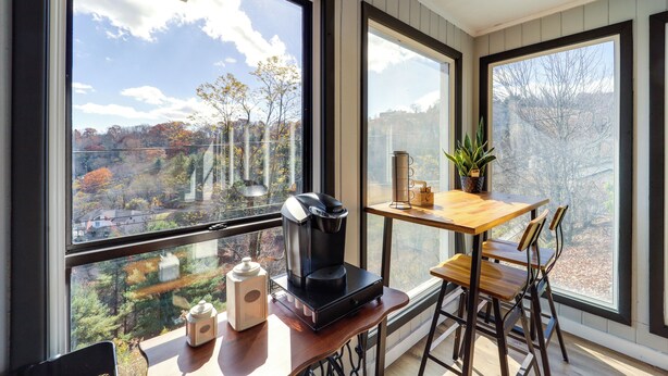 A room with large windows offering a view of a forested area. A coffee maker is on a wooden table, and there are chairs around it. - Pet-friendly Boone Home w/ Scenic Mountain Views! (Boone)
