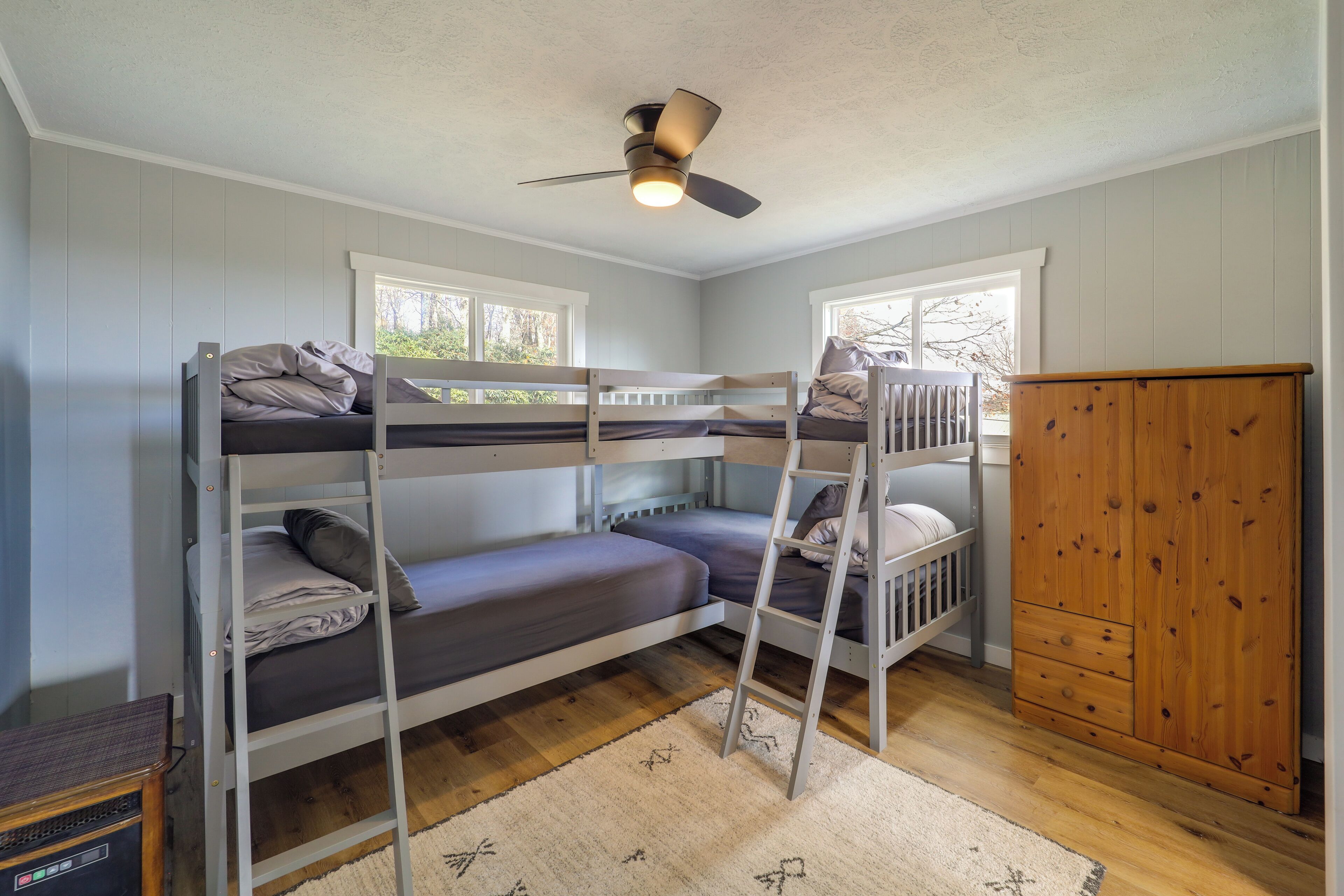 A bunk bed room with a ceiling fan, wooden wardrobe, and a window with a view of trees.