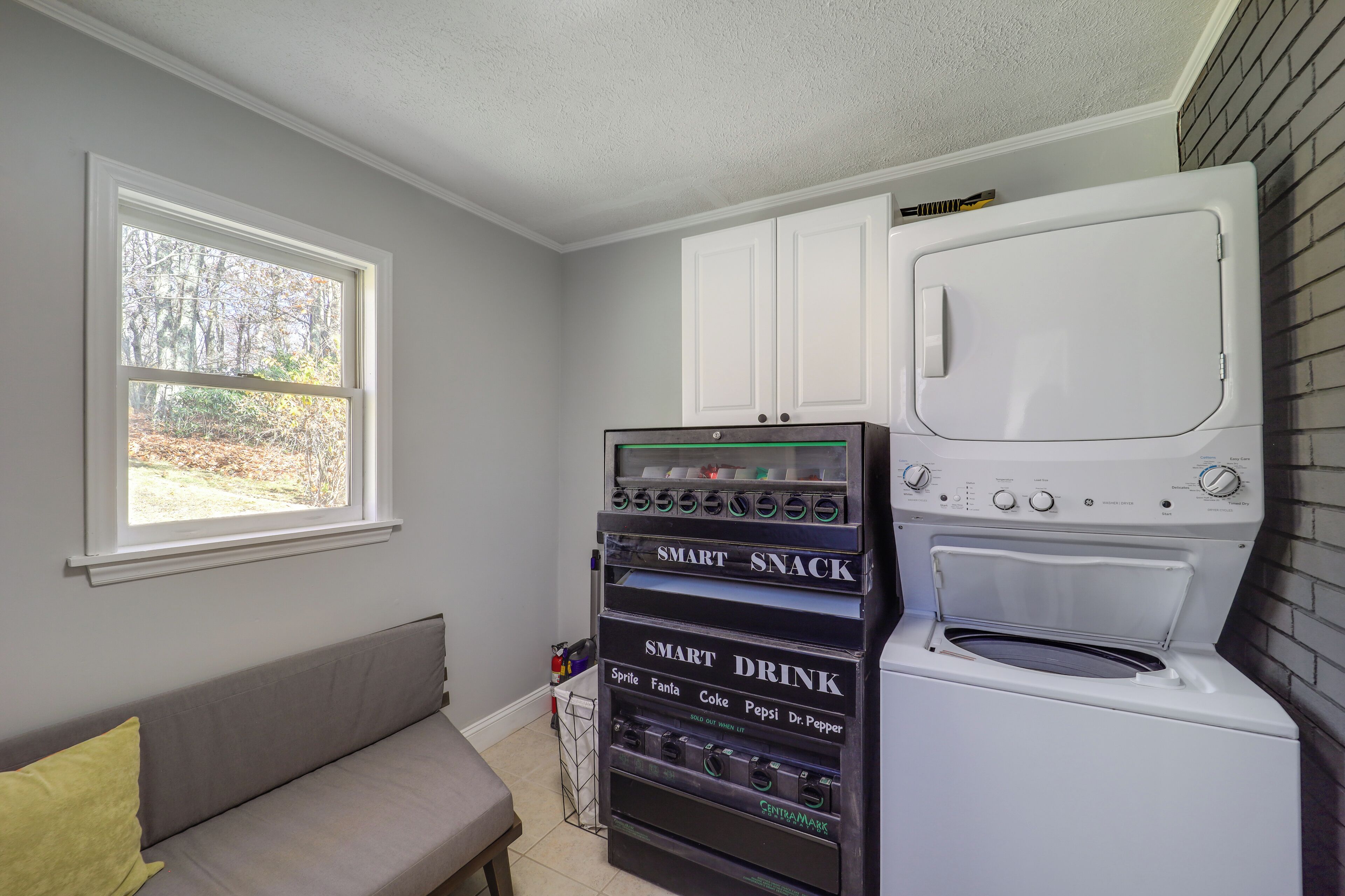 A laundry room with a washer and dryer, a vending machine with snacks and drinks, and a window with a view of trees.