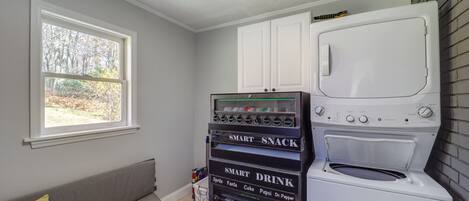 A laundry room with a washer and dryer, a vending machine with snacks and drinks, and a window with a view of trees.