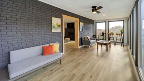A modern living room with a grey brick wall, a grey sofa with orange cushions, a wooden coffee table, and a dining area with a high table and chairs. - Pet-friendly Boone Home w/ Scenic Mountain Views! (Boone)
