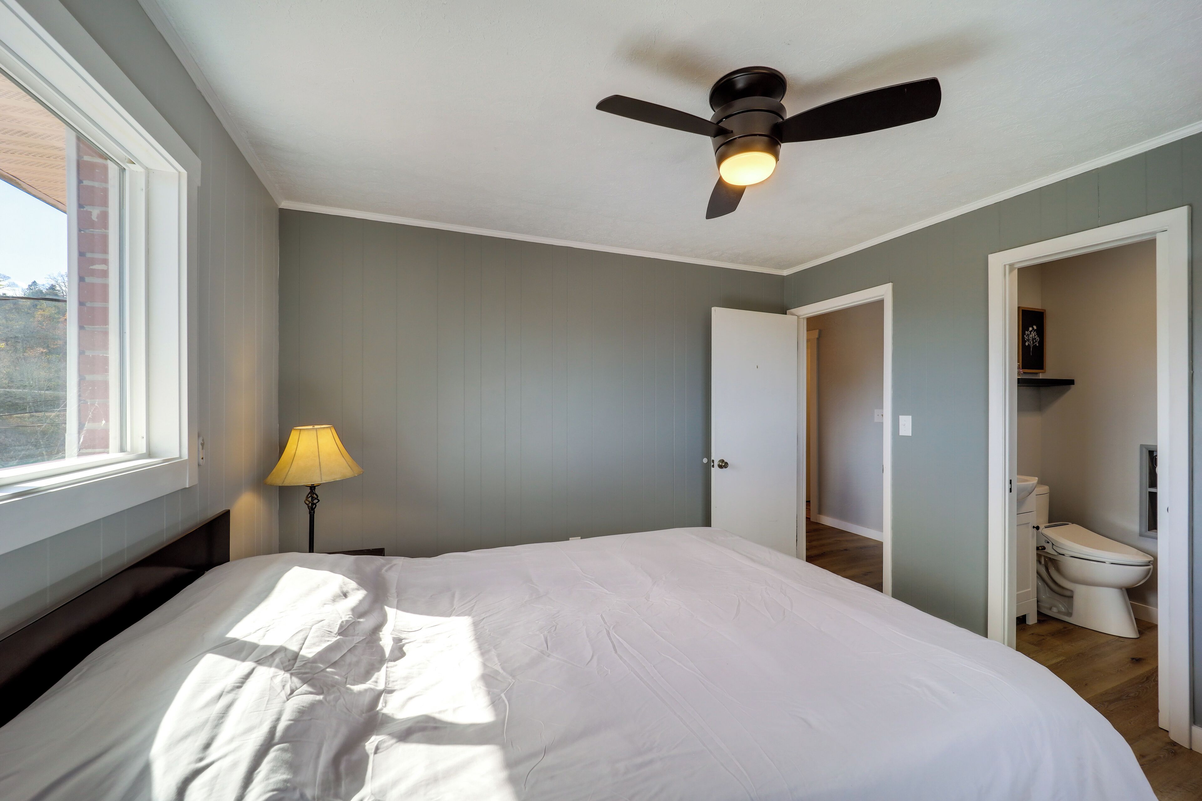 A bedroom with a bed, a ceiling fan, a window, and a door leading to a bathroom.