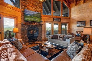 L’intérieur chaleureux d’un chalet avec une cheminée en pierre, des canapés en cuir et une grande fenêtre donnant sur un paysage magnifique.