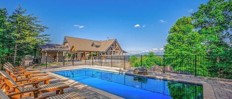 A swimming pool with wooden deck chairs, a covered seating area, and a building with a gabled roof in the background.