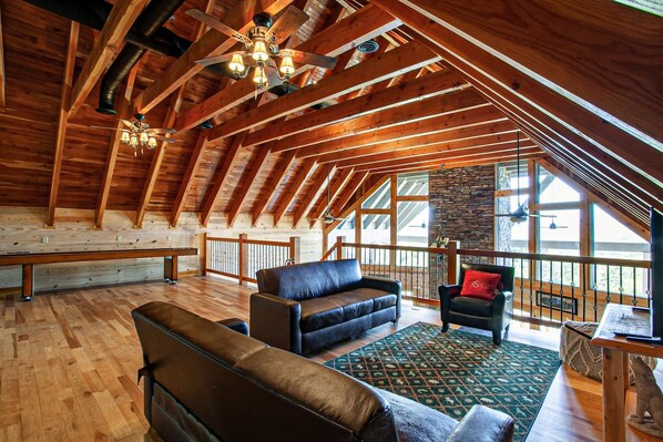 A spacious loft with high ceilings, exposed wooden beams, a large seating area with leather sofas, a stone accent wall, and large windows offering views.