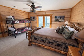 Une chambre dans un chalet en bois, avec un lit superposé, un grand lit doté d’une tête de lit sur le thème de la forêt et un ventilateur de plafond.