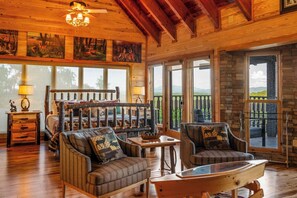 A cozy cabin interior with a rustic wooden ceiling, a large bed with a wooden headboard, two upholstered armchairs, a glass coffee table, and a dining table.