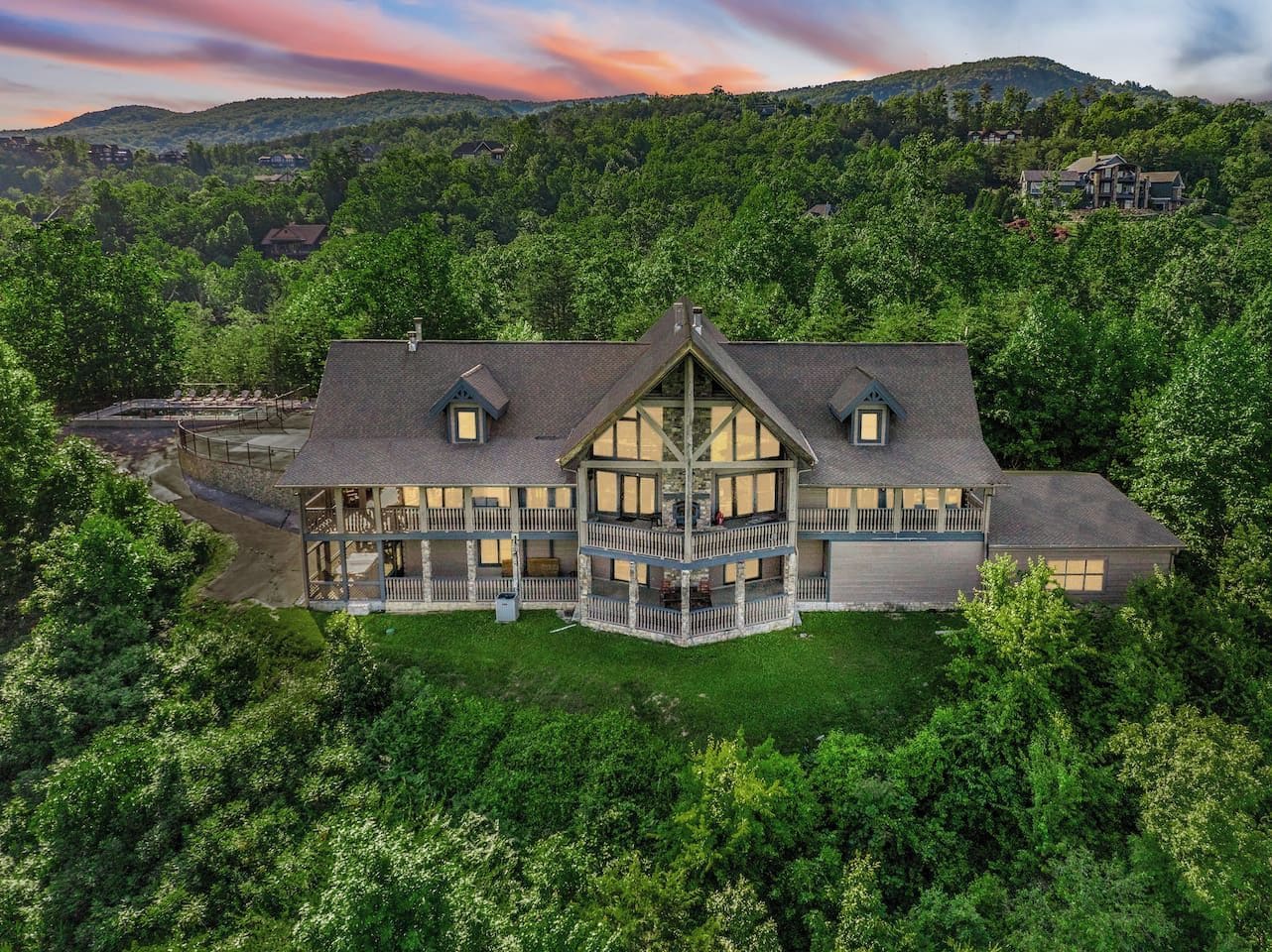 A large, multi-story house with a prominent balcony, surrounded by dense greenery and hills in the background.