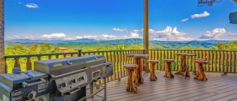 Una terraza exterior con una parrilla y taburetes de madera, con vistas a un paisaje montañoso.