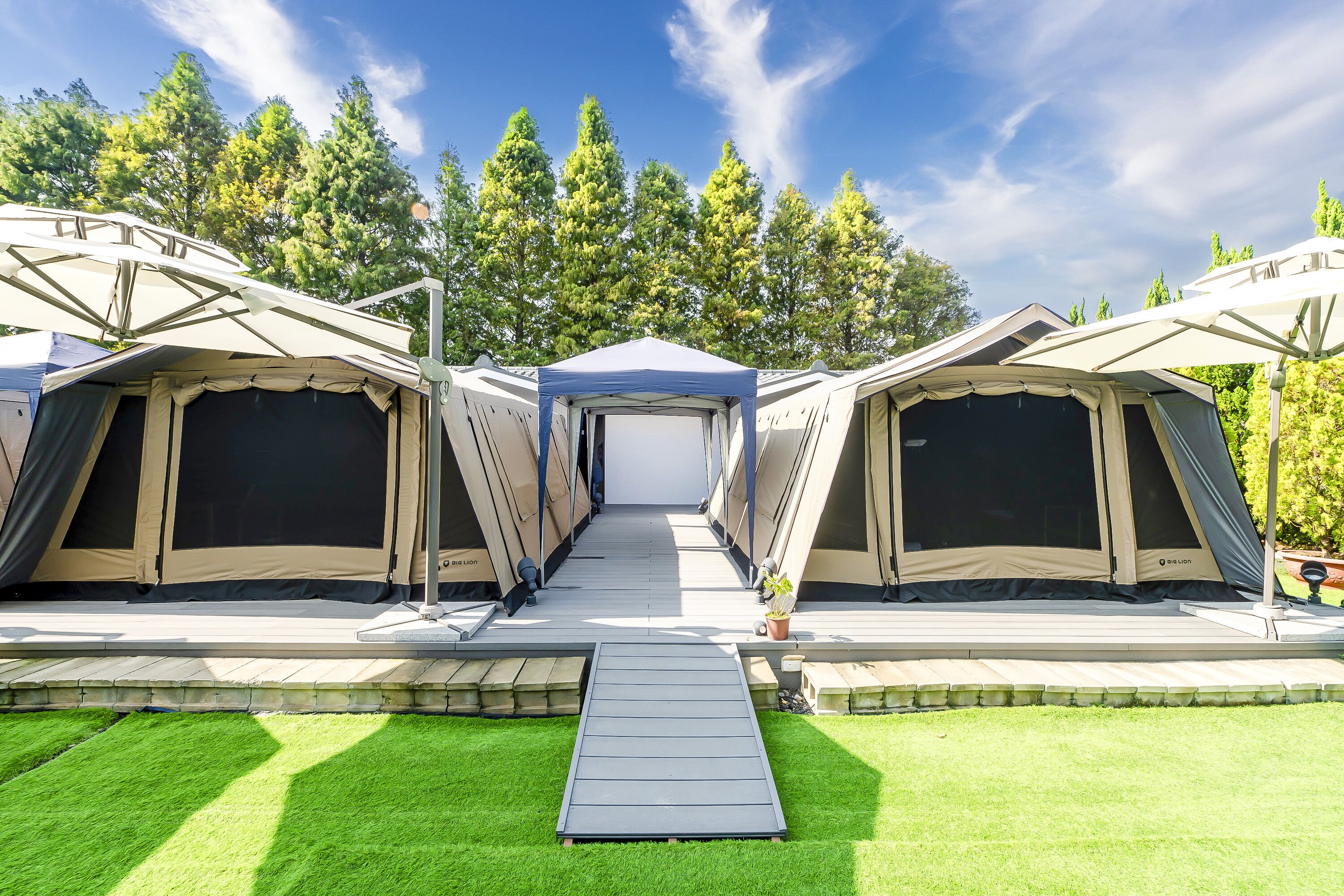 A row of camping tents with a central walkway leading to a covered area, surrounded by a grassy area and trees.