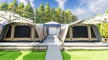 A row of camping tents with a central walkway leading to a covered area, surrounded by a grassy area and trees.