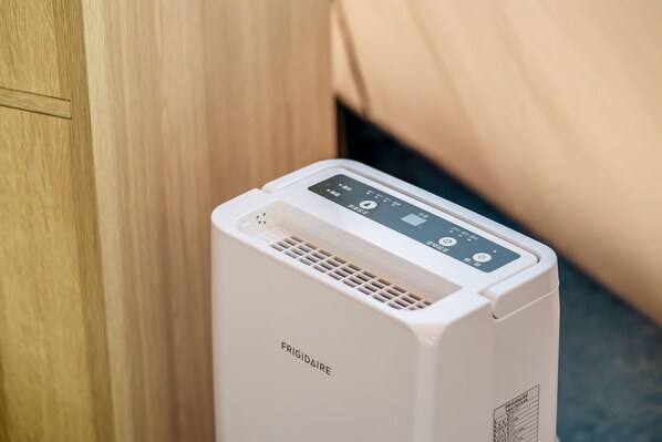 A white portable air conditioner with a digital display and control panel, placed against a wooden cabinet and a beige fabric backdrop.