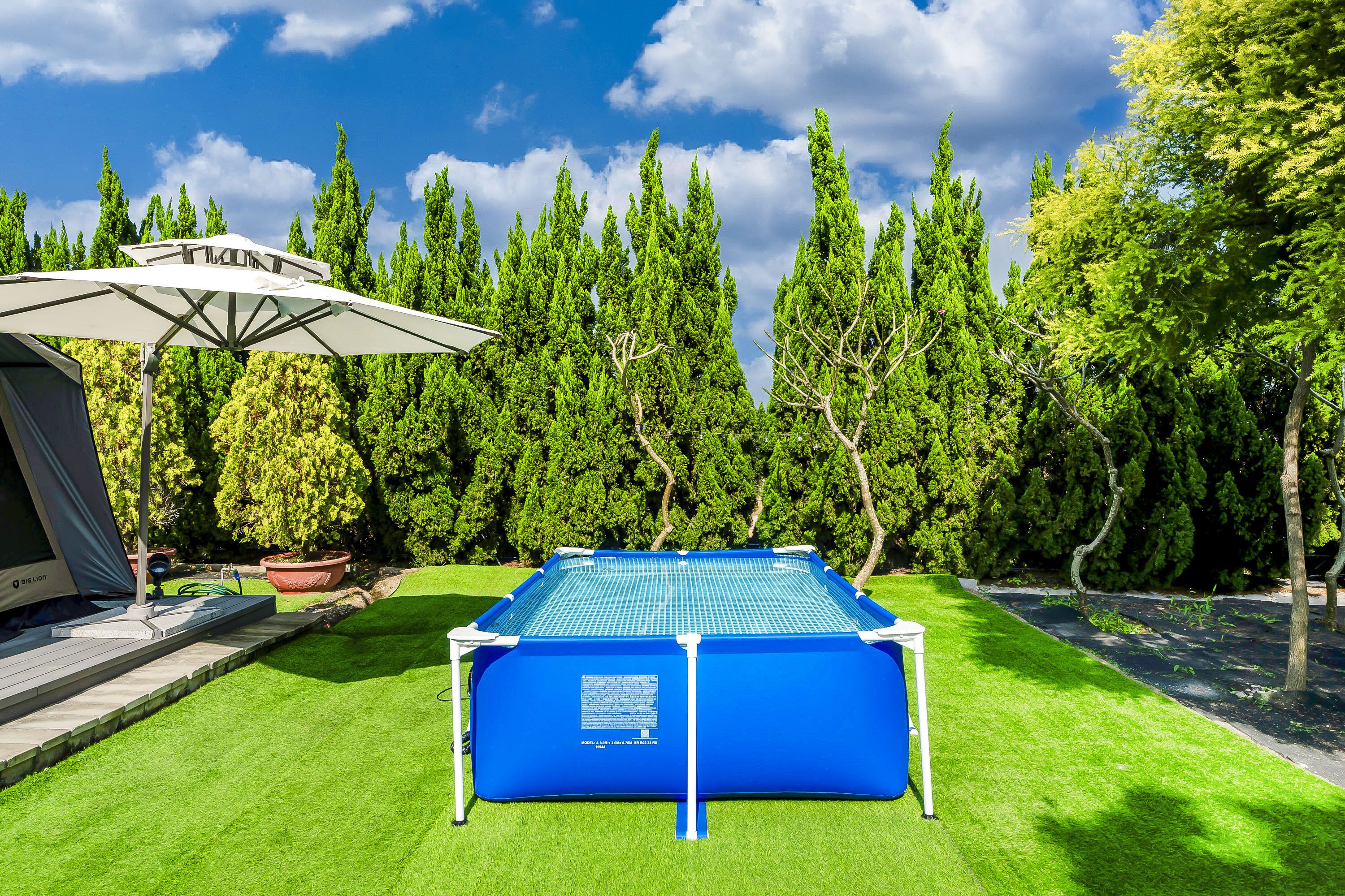 Una piscina con cubierta azul, rodeada de césped cuidado y altos árboles.