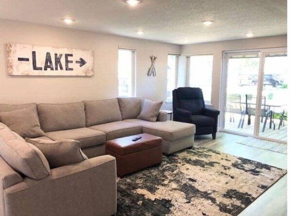 A living room with a beige sectional sofa, a brown ottoman, and a black and white rug. There is a wall-mounted sign that reads "LAKE" with an arrow pointing to the right. - Completely remodeled home within walking distance to the lake! (Clear Lake)