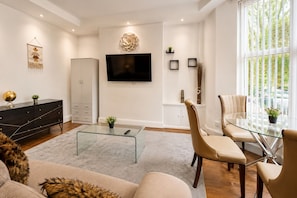A modern living room with a flat-screen TV, a glass coffee table, a sofa, and a dining area with a glass table and chairs. - Botanic Gardens Oasis: Urban Convenience & Comfort (Liverpool)