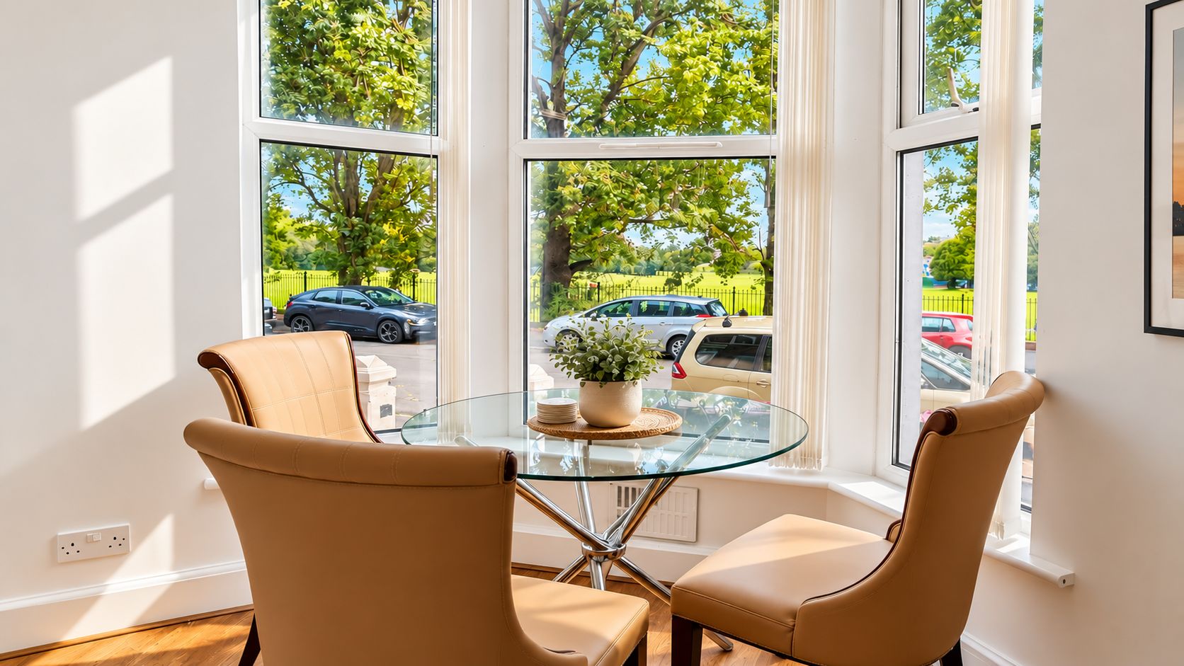 A room with a glass table and two chairs, a potted plant, and a view of a parking lot through large windows.